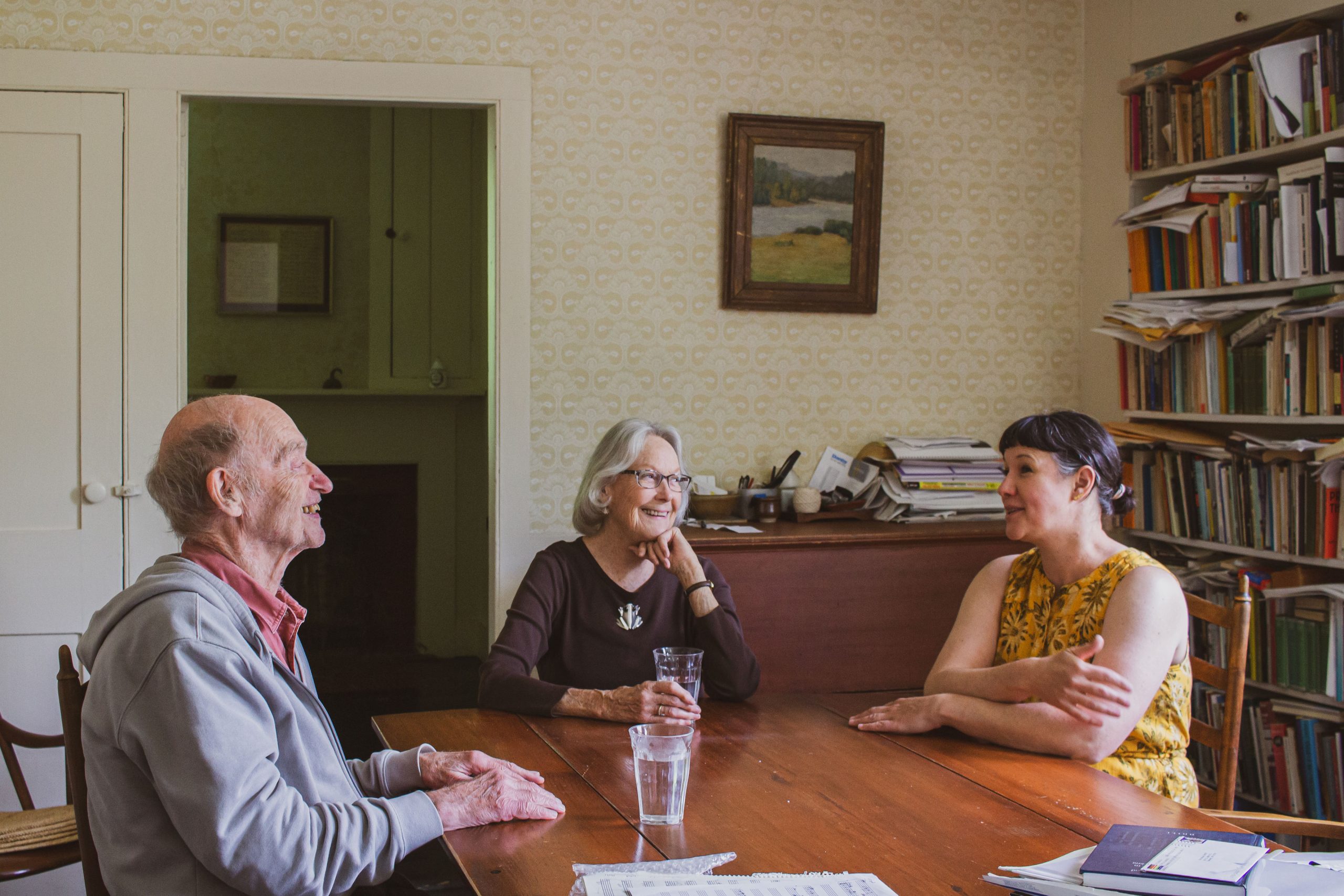 3 people seated at at table, white male on the left in his 90s, hands on table, center white woman in her 80s hand on chin looking toward white woman in her 40s, smiling, dark hair and yellow jumpsuit, arms on the table.
